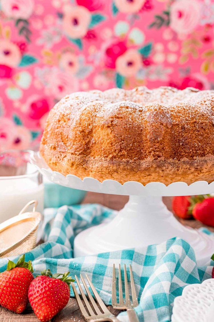 Whipping cream cake on a white cake stand on a blue gingham napkin with a pink floral background.