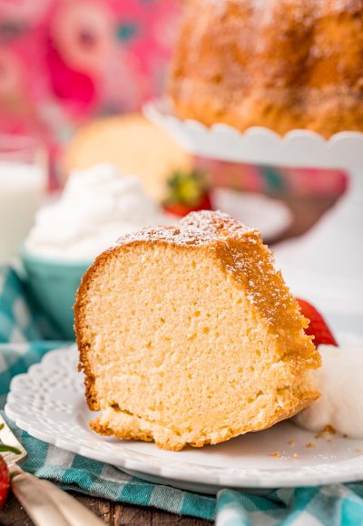 Close up photo of a slice of whipping cream cake on a white plate with whipped cream and a strawberry on the side.