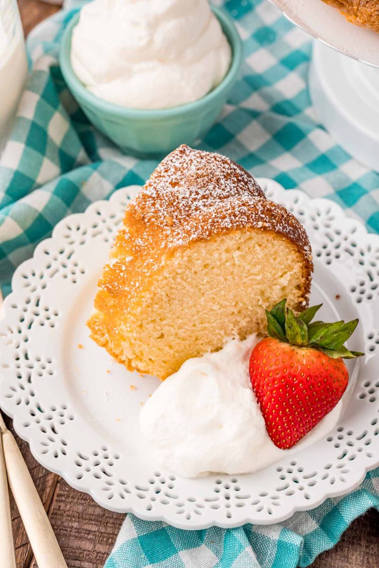 A slice of whipping cream cake on a white plate with whipped cream and a strawberry on a teal gingham napkin.