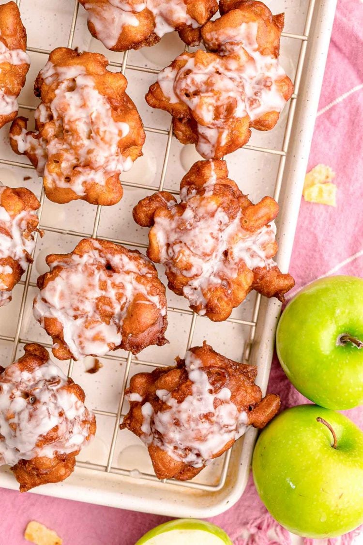 Overhead photo of apple fritters on a baking sheet on a pink napkin with apples on the side.
