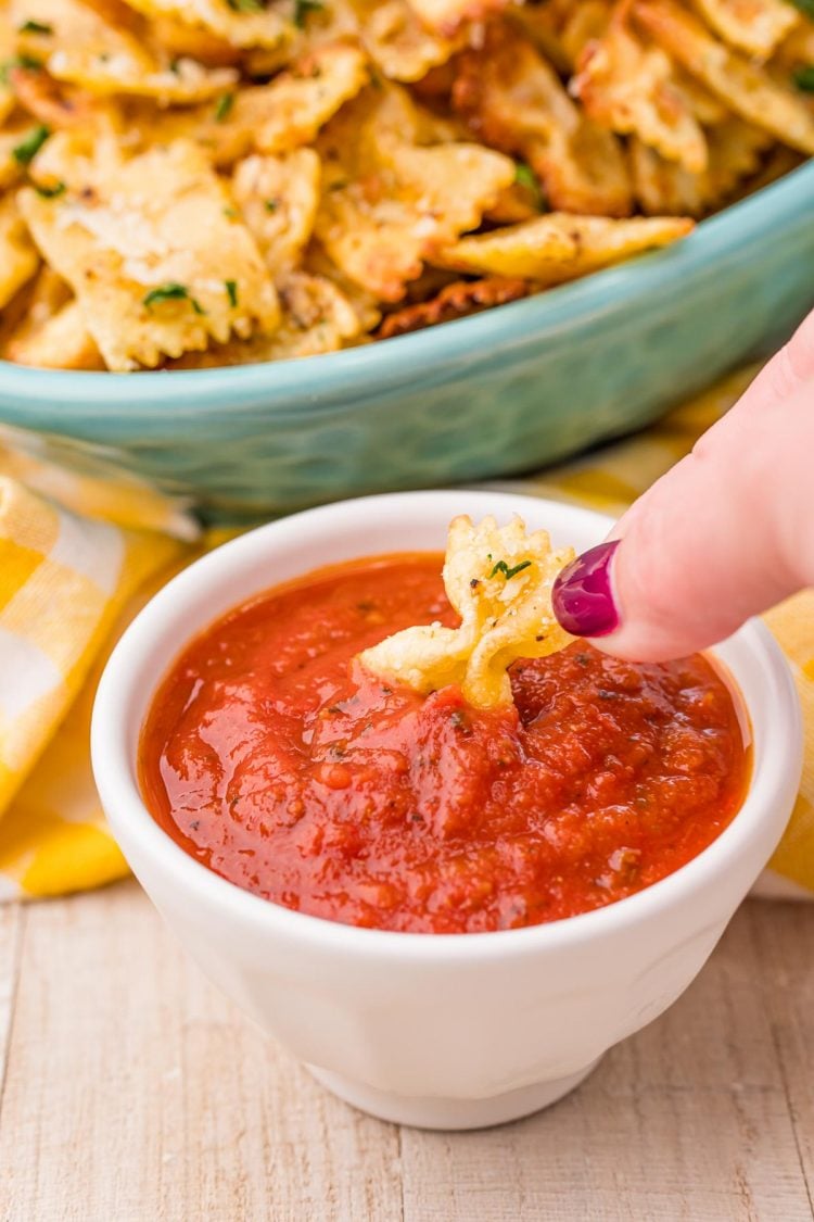 A woman's hand dipping a pasta chip into a small white bowl filled with marinara sauce.