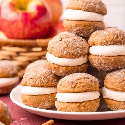 Close up photo of a stack of mini apple cider whoopie pies on a white plate with apples in the background.