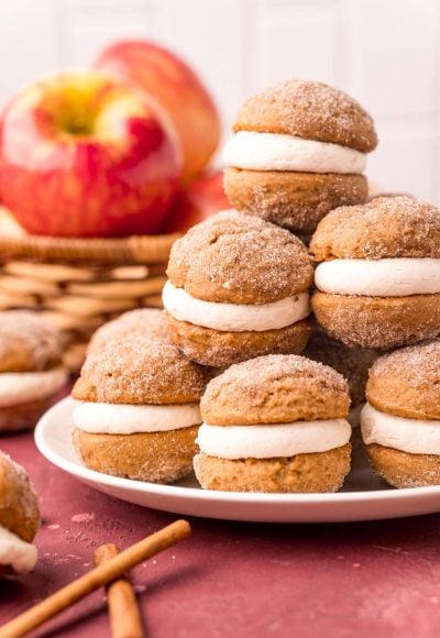Close up photo of a stack of mini apple cider whoopie pies on a white plate with apples in the background.