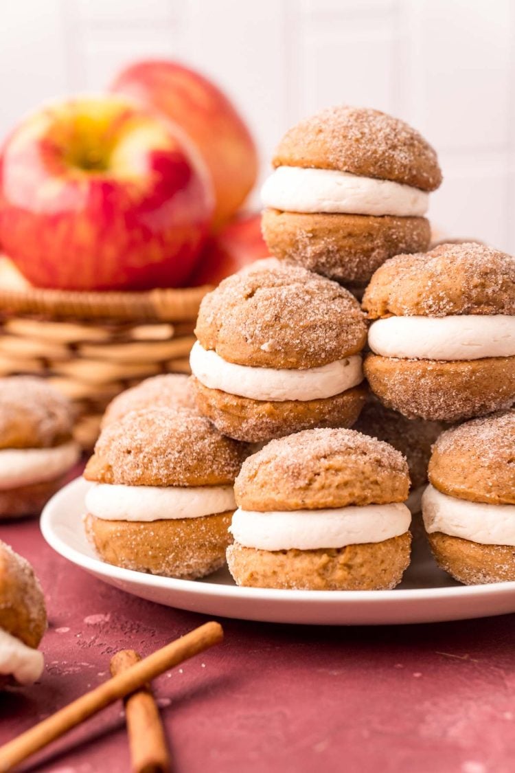 Close up photo of a stack of mini apple cider whoopie pies on a white plate with apples in the background.