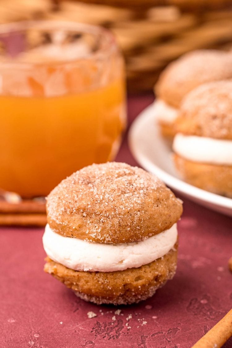 Close up photo of an apple cider whoopie pie on a maroon surface with a glass of apple cider in the background.