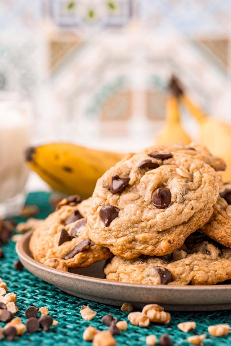 Banana bread cookies on a small plate on a teal placemat with bananas in the background.