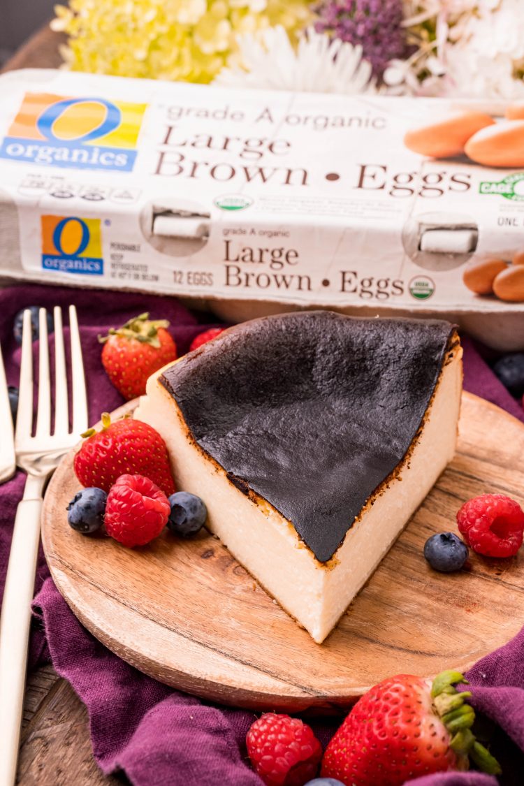 Close up photo of a slice of basque cheesecake on a wooden plate with fresh berries and gold forks. A carton of eggs is in he background.