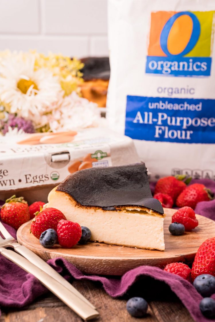 A slice of basque cheesecake on a wooden plate on a purple napkin with fresh berries around and a bag of flour in the background.