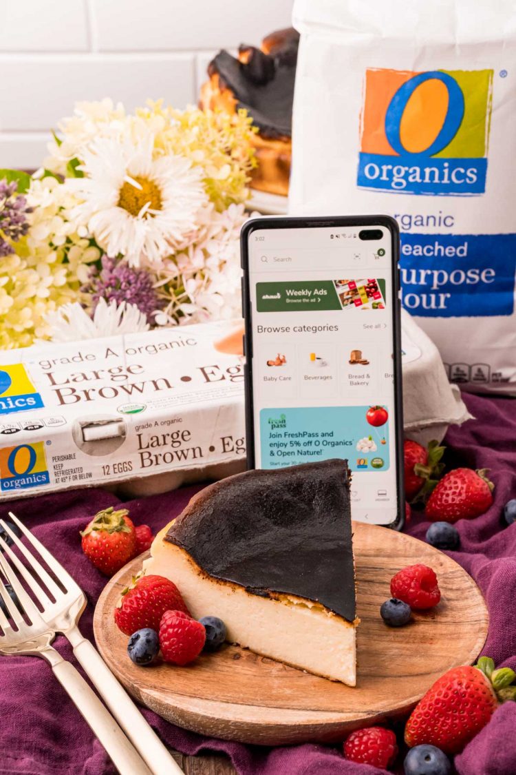 Close up photo of a slice of burnt cheesecake on a wooden plate with a carton of eggs and bag of flour in the background with a phone propped against them with the Shaw's for U app open.