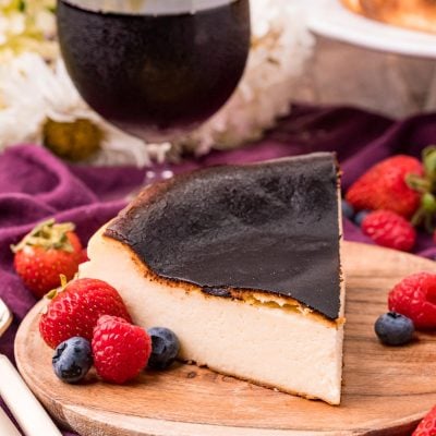 Close up photo of Basque Burnt Cheesecake on a wooden plate with fresh berries scattered around and a glass of red wine in the background.