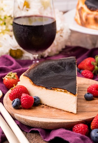 Close up photo of Basque Burnt Cheesecake on a wooden plate with fresh berries scattered around and a glass of red wine in the background.