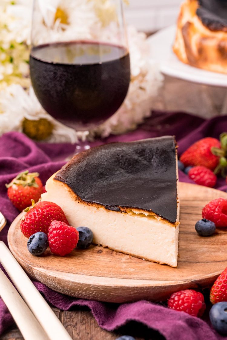 Close up photo of Basque Burnt Cheesecake on a wooden plate with fresh berries scattered around and a glass of red wine in the background.