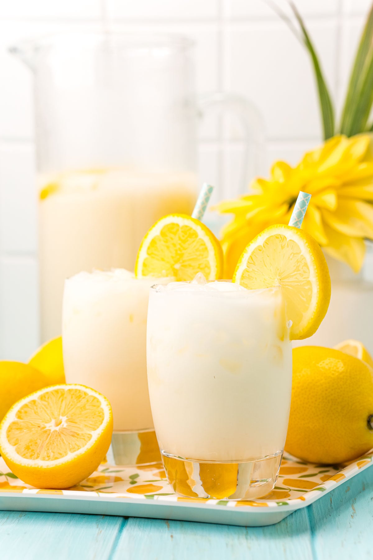 Straight on photo of two glasses of creamy lemonade on a tray with lemons and a pitcher in the background.