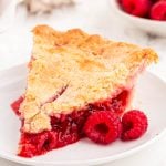 Close up photo of a slice of raspberry pie on a white plate with fresh raspberries around it.