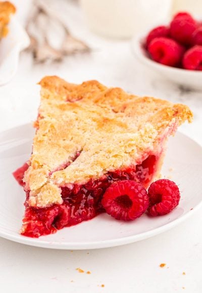 Close up photo of a slice of raspberry pie on a white plate with fresh raspberries around it.