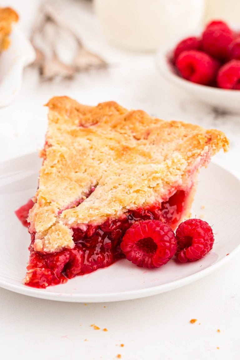 Close up photo of a slice of raspberry pie on a white plate with fresh raspberries around it.