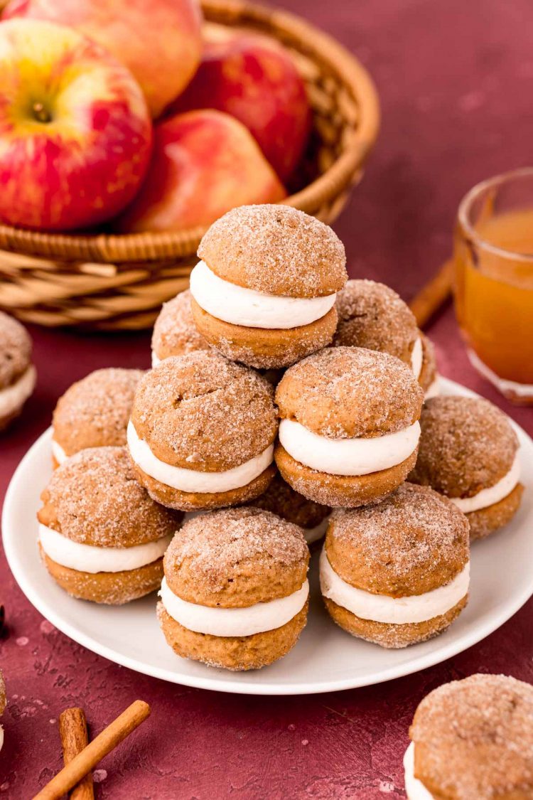 Mini apple cider whoopie pies stacked on a white plate on a maroon surface with apples in the background.