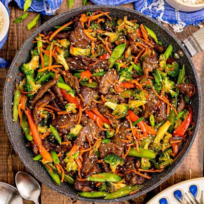 Overhead photo of a pan with beef stir fry in it on a wooden table with blue napkin next to it.