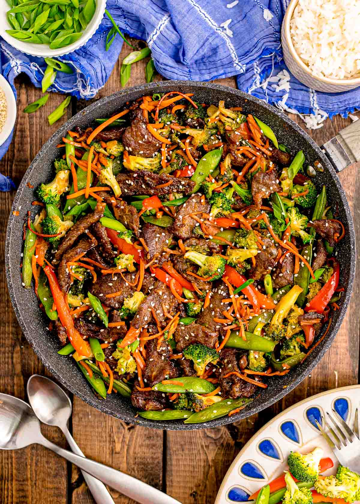 Overhead photo of a pan with beef stir fry in it on a wooden table with blue napkin next to it.