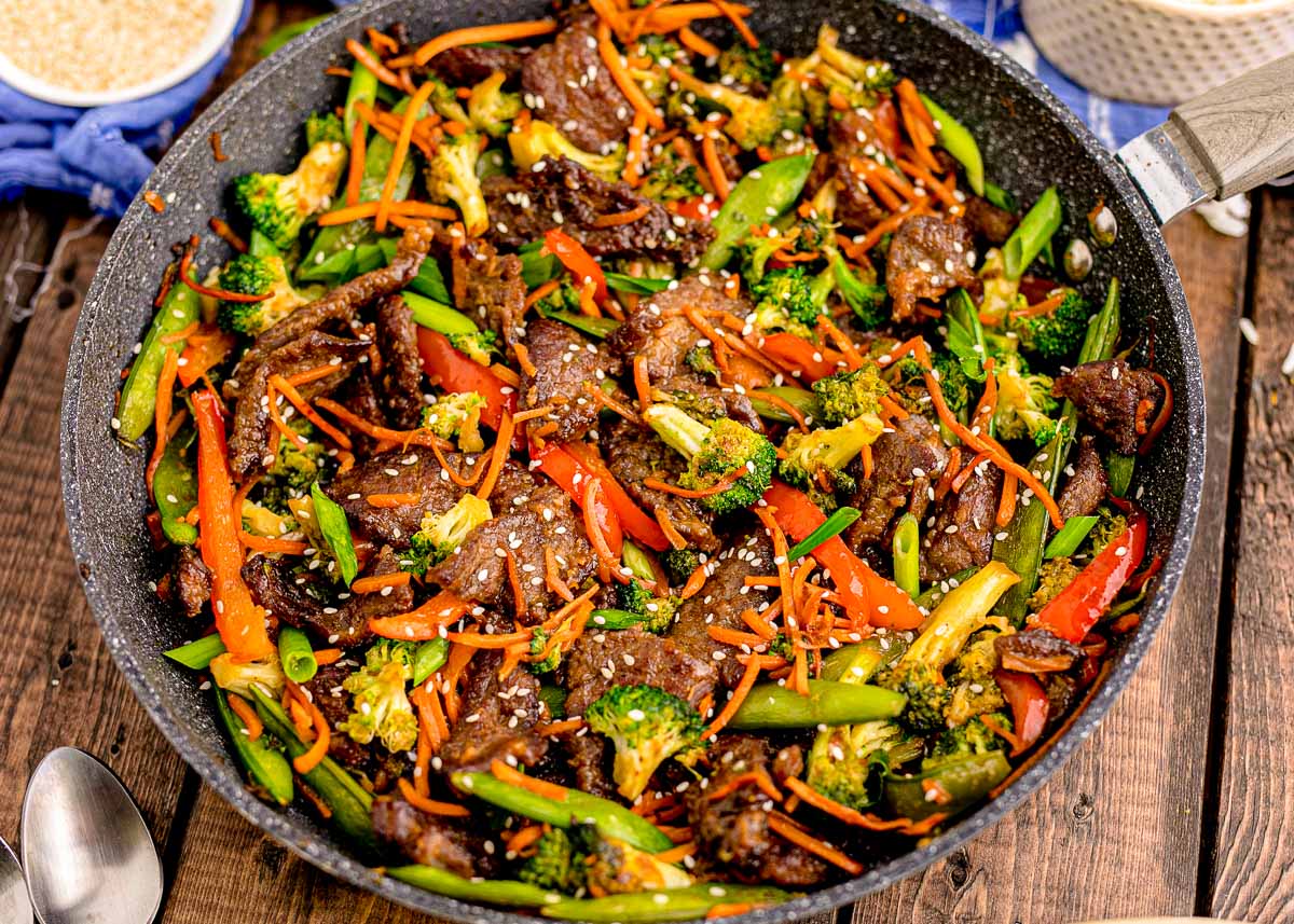 Overhead photo of a pan with beef stir fry in it on a wooden table with blue napkin next to it.