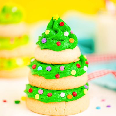 Close up photo of a Christmas tree cookie made with stacked sugar cookies and green frosting on a white surface with a yellow background.