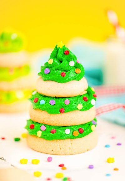 Close up photo of a Christmas tree cookie made with stacked sugar cookies and green frosting on a white surface with a yellow background.