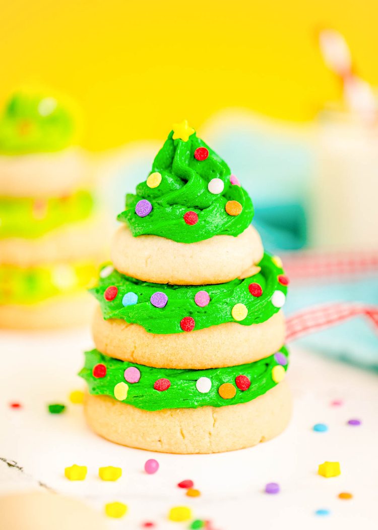 Close up photo of a Christmas tree cookie made with stacked sugar cookies and green frosting on a white surface with a yellow background.