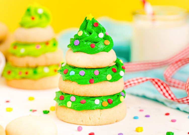 Close up photo of a Christmas tree cookie made with stacked sugar cookies and green frosting on a white surface with a yellow background.