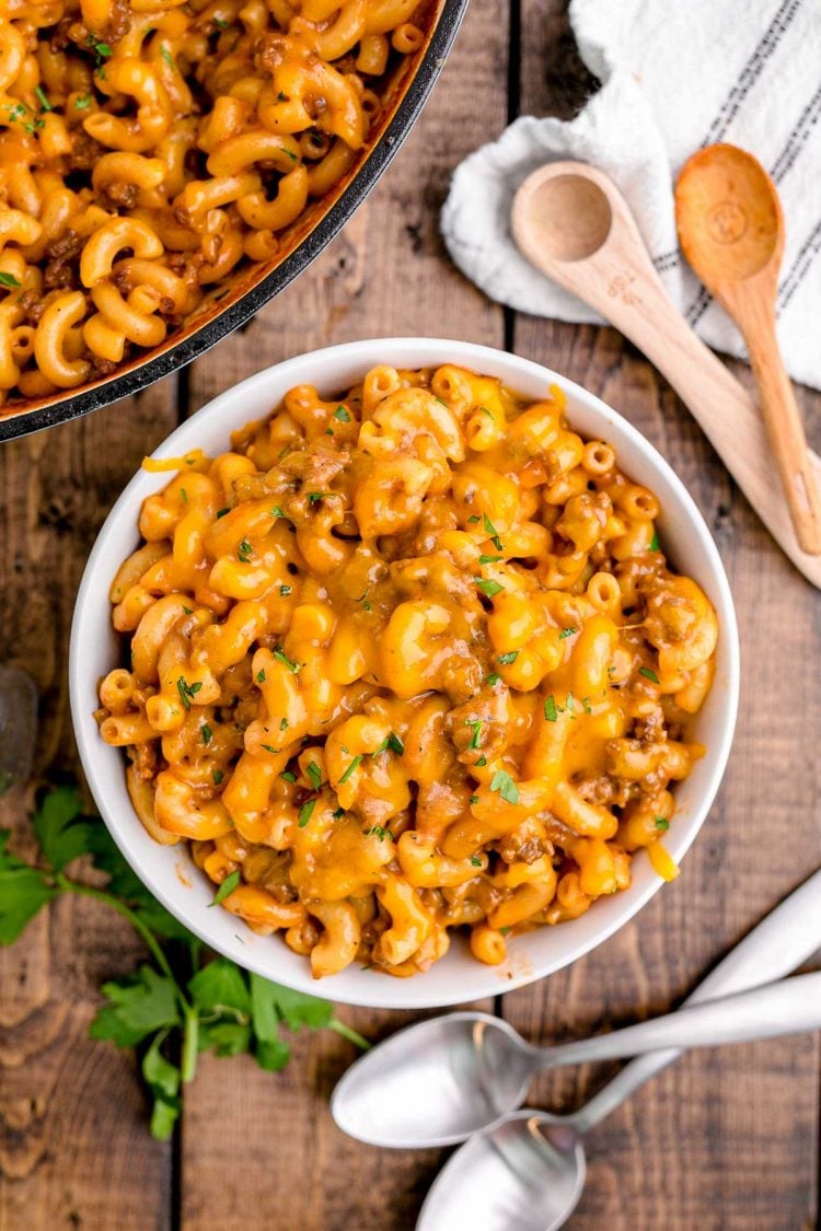 Close up photo of a white bowl with homemade hamburger helper on a wooden table with spoons and linens around it.