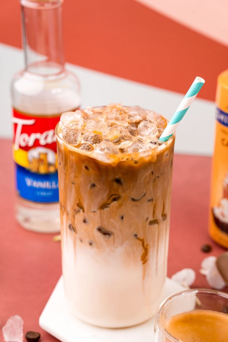 A close up photo of a glass with iced caramel macchiato in it and a bottle of caramel syrup and vanilla simple syrup in the background.