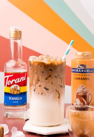 Close up photo of an iced caramel macchiato on a white marble coaster on a dark red table with caramel sauce and vanilla simple syrup bottles in the background.