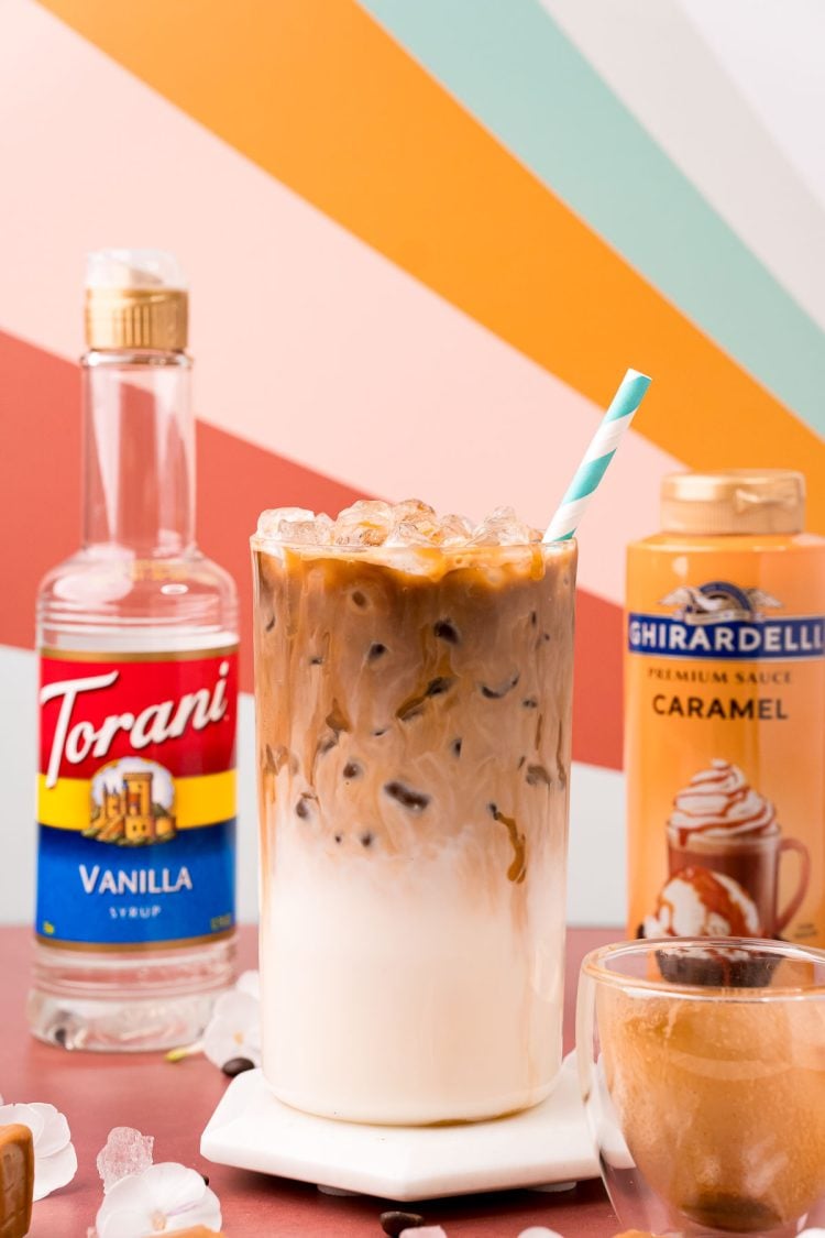 Close up photo of an iced caramel macchiato on a white marble coaster on a dark red table with caramel sauce and vanilla simple syrup bottles in the background.
