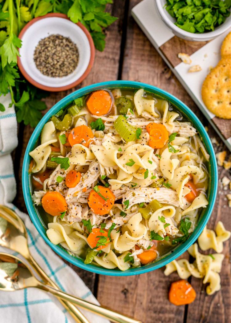 Overhead photo of chicken noodle soup in a teal bowl on a wooden table with herbs and a napkin around it.