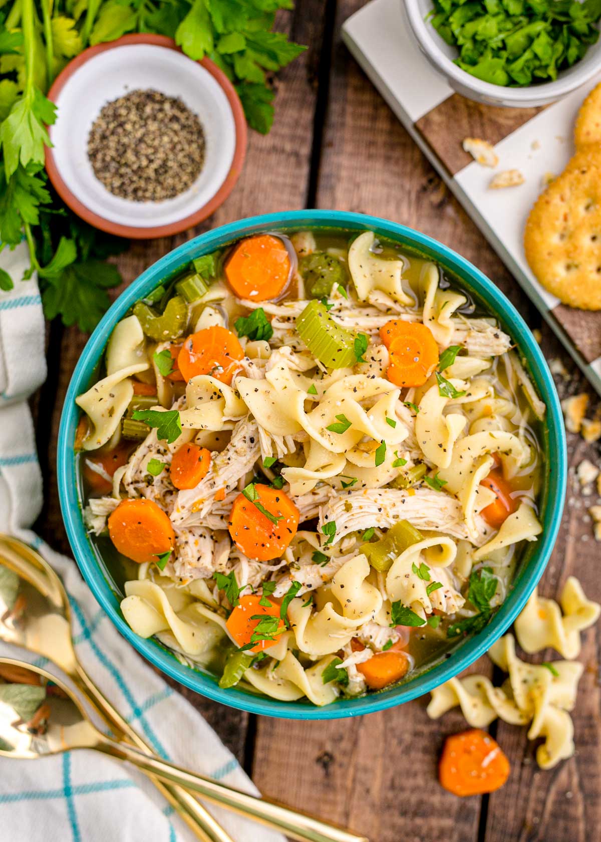 Overhead photo of chicken noodle soup in a teal bowl on a wooden table with herbs and a napkin around it.