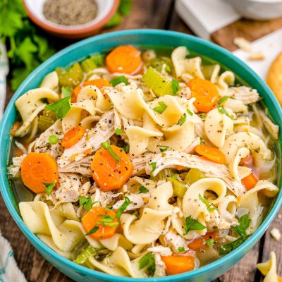 Close up photo of a teal bowl filled with chicken noodle soup with herbs and spices in the background.