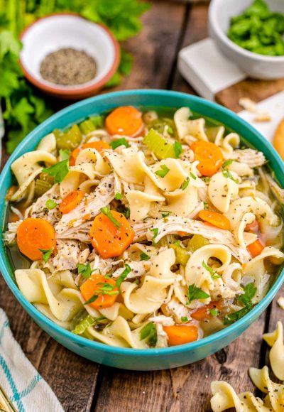 Close up photo of a teal bowl filled with chicken noodle soup with herbs and spices in the background.