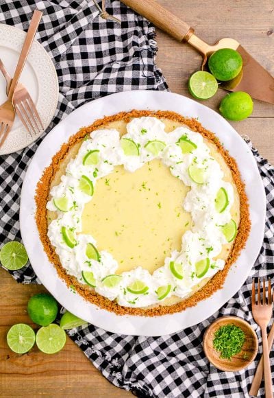 Overhead photo of a key lime pie in a white pie dish on a black and white gingham napkin on a wooden table with lime scattered around.