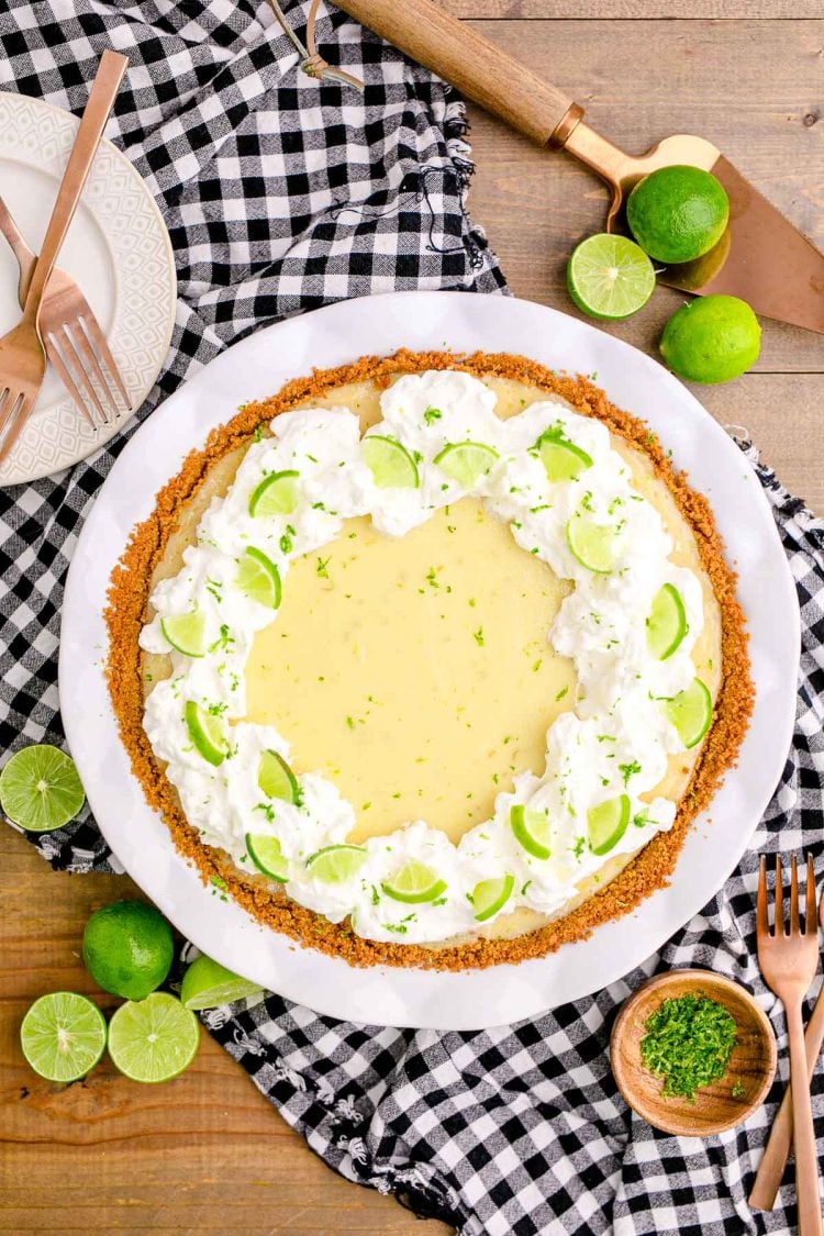 Overhead photo of a key lime pie in a white pie dish on a black and white gingham napkin on a wooden table with lime scattered around.