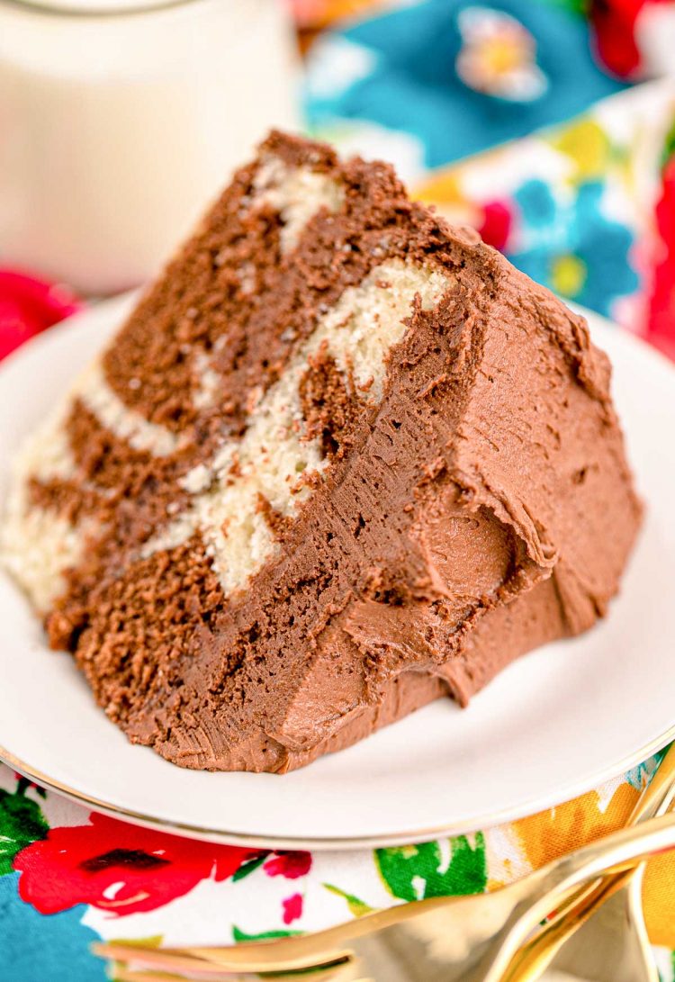 Close up photo of a slice of marble cake on a white plate with a glass of milk and gold fork next to it on a floral napkin.