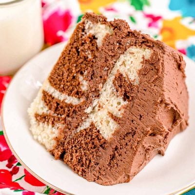 Close up photo of a slice of marble cake on a white plate with a glass of milk and gold fork next to it on a floral napkin.