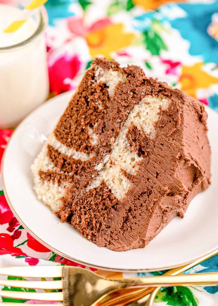 Close up photo of a slice of marble cake on a white plate with a glass of milk and gold fork next to it on a floral napkin.