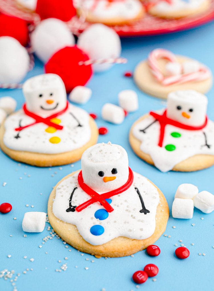 Melted Snowman Cookies on a blue surface with candy and marshmallows scattered around.