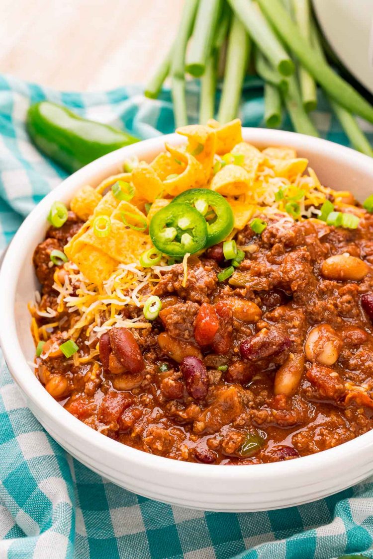 Close up photo of chili in a white bowl on a gigham napkin with peppers in the background.