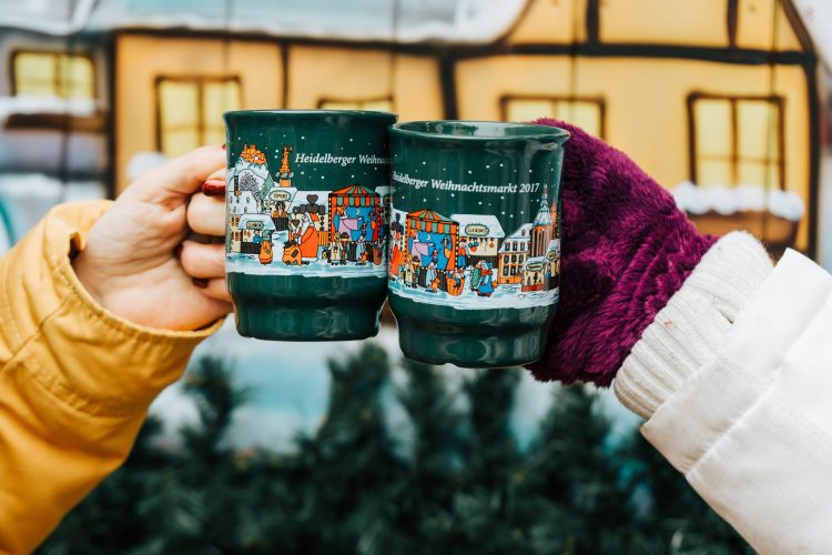 Two women's mittened hands clink mugs of kinder punch at a German Christmas Market.