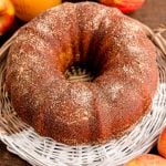 A whole apple cider donut bundt cake on a wicker tray with apples and apple cider in the background.