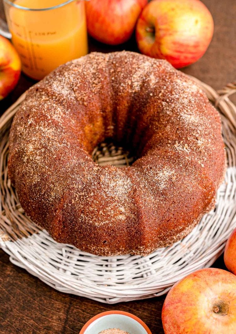 A whole apple cider donut bundt cake on a wicker tray with apples and apple cider in the background.
