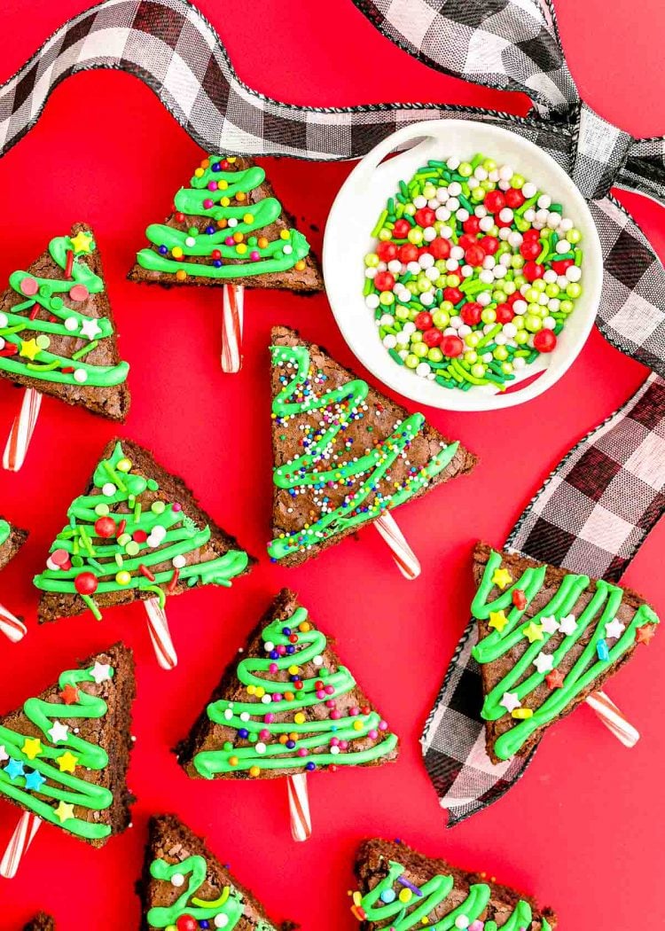 Overhead photo of brownie Chrsitmas trees on a red surface with holiday ribbon and sprinkles around them.
