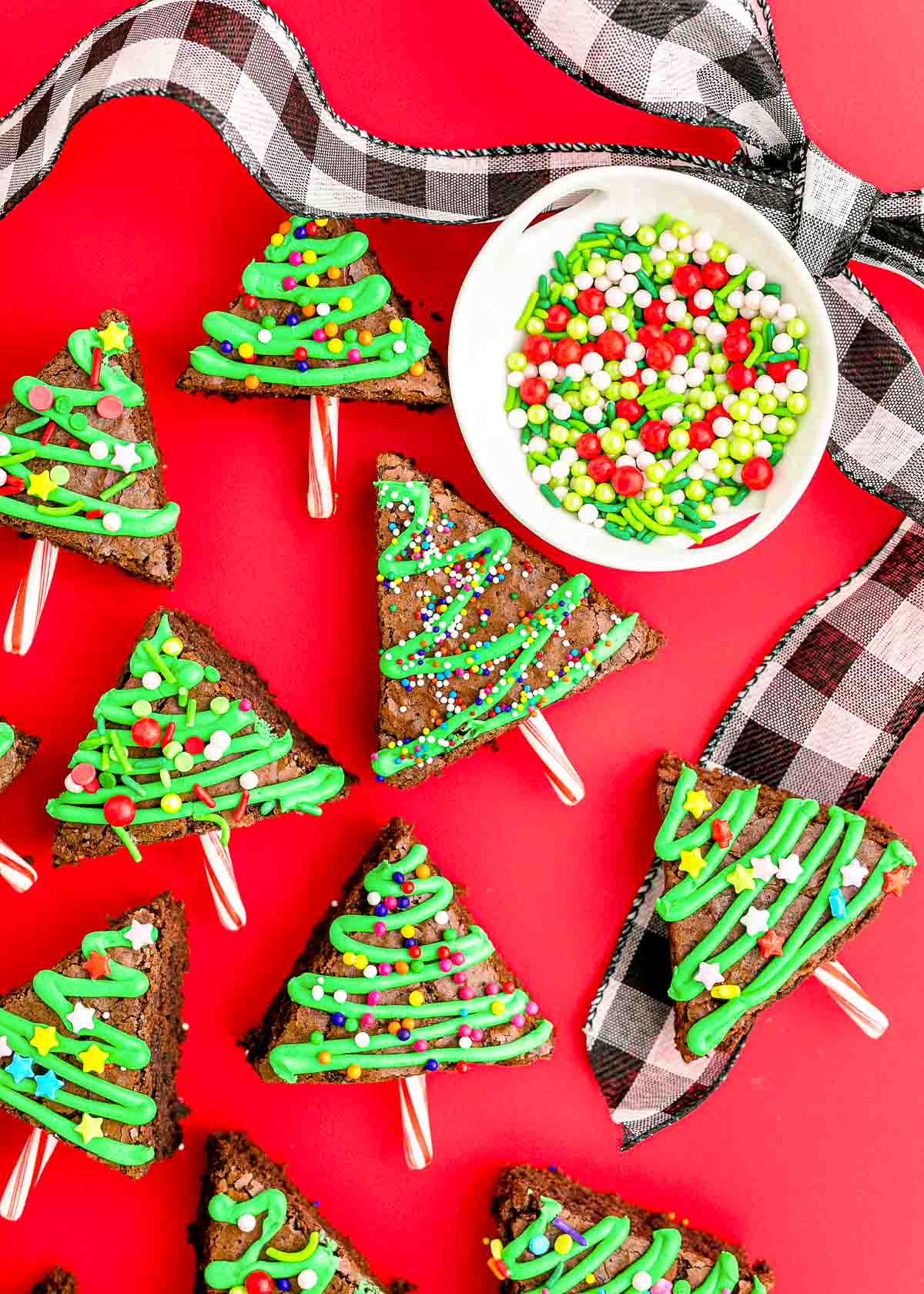 Overhead photo of brownie Chrsitmas trees on a red surface with holiday ribbon and sprinkles around them.