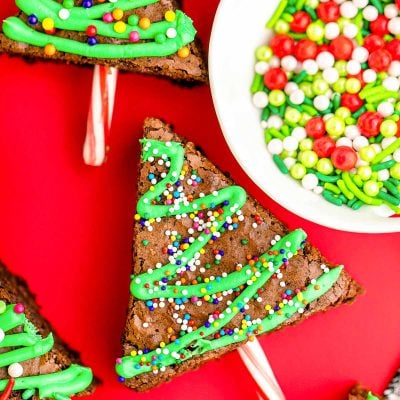 Close up photo of brownie christmas trees laying on a red surface with a bowl of sprinkles next to them.