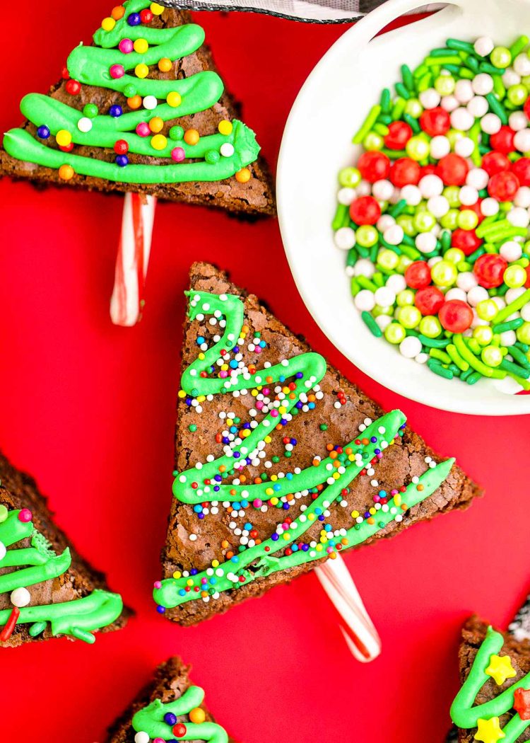 Close up photo of brownie christmas trees laying on a red surface with a bowl of sprinkles next to them.